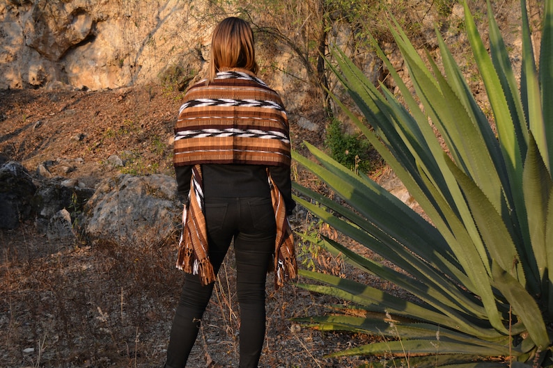 May include: A woman wearing a brown, white and black woven shawl with fringe. The shawl has a geometric pattern. She is standing in front of a large green plant with long, sharp leaves.