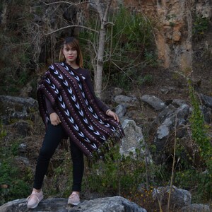 May include: A woman wearing a brown and white patterned poncho with black trousers and pink shoes stands on a large rock in a rocky, outdoor setting.