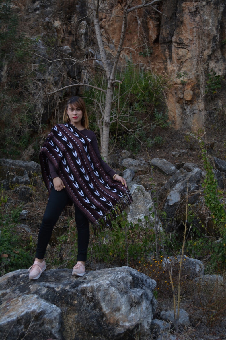 May include: A woman wearing a brown and white patterned poncho with black trousers and pink shoes stands on a large rock in a rocky, outdoor setting.