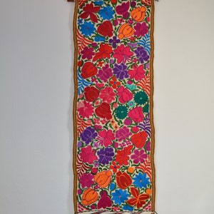 May include: A white tapestry with a colorful floral pattern. The tapestry is embroidered with red, pink, orange, yellow, blue, green, and purple flowers. The tapestry is hanging on a white wall.