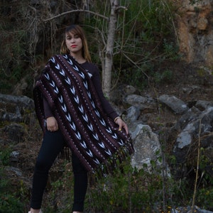 May include: A woman wearing a brown and white patterned poncho with black leggings and pink trainers. She is standing on a large rock in a rocky, green, and brown outdoor setting.