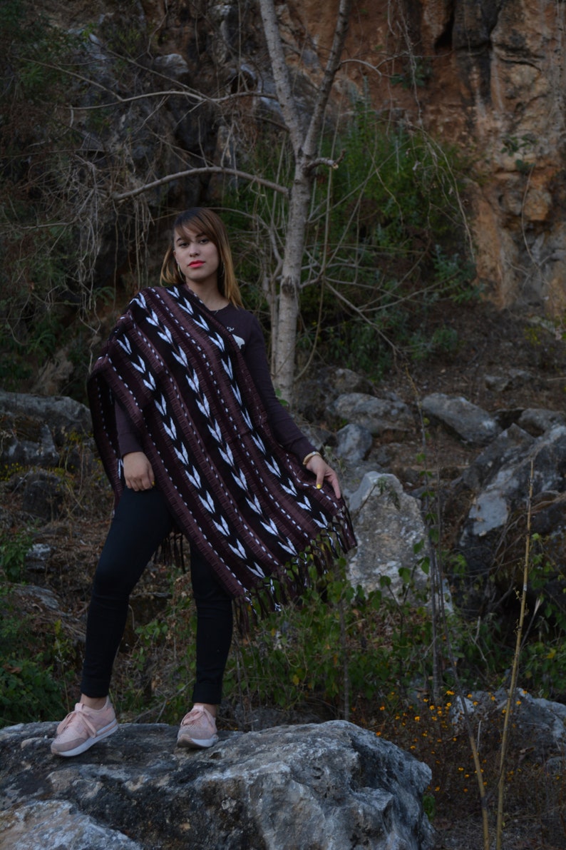 May include: A woman wearing a brown and white patterned poncho with black leggings and pink trainers. She is standing on a large rock in a rocky, green, and brown outdoor setting.