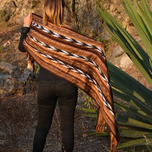 May include: A person wearing black jeans and a brown, black and white striped shawl with tassels. The shawl is draped over the person's shoulders and is flowing in the wind. The person is standing in a field with green plants in the background.