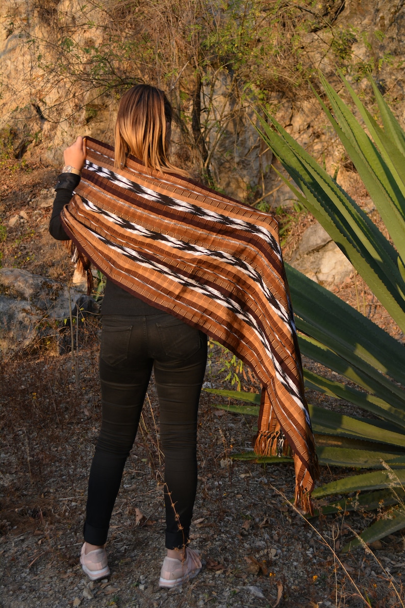 May include: A person wearing black jeans and a brown, black and white striped shawl with tassels. The shawl is draped over the person's shoulders and is flowing in the wind. The person is standing in a field with green plants in the background.
