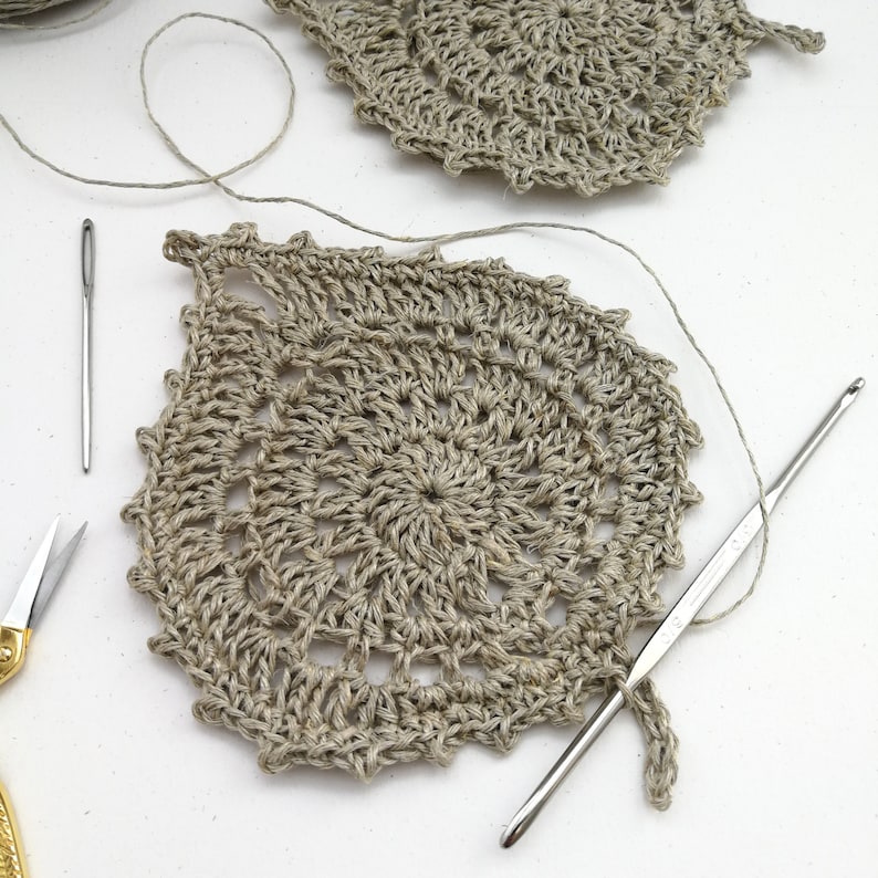 A set of leaf shaped coasters are sat on a white background. They have been crocheted in the round showing a small circle in the middle which expands out into a leaf shape. It has picots all the way around and a small stem at the bottom.