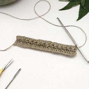 Two rows of crochet work in two different stitches with a silver hook still attached. The colour of the yarn is warm grey and work looks textured.