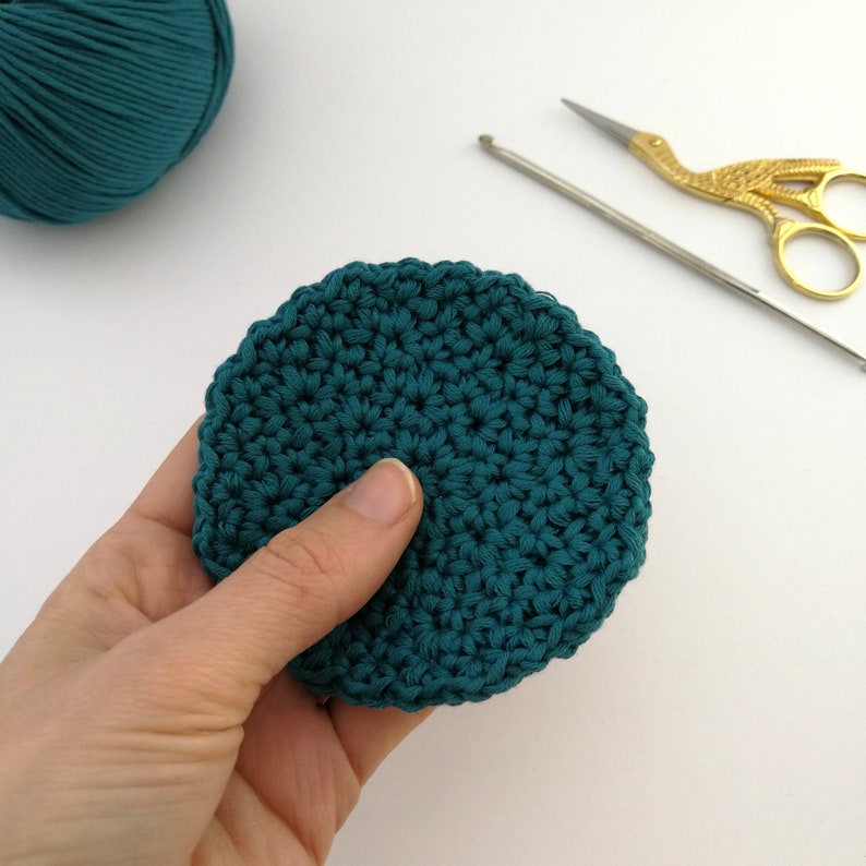A left hand is holding one teal face scrubbie with the thumb at the centre of the round. The background is off white and there is a silver hook and pair of scissors in the top right.