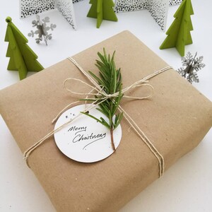 A Christmas present is wrapped in brown paper and has a circular white tag with "Merry Christmas" written on it in black ink. It is secured with hemp string tied into a bow in the middle and a sprig of rosemary placed underneath it.