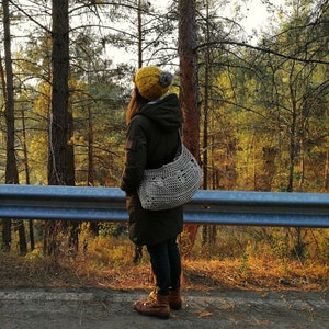 A model is stood at the side of the road looking out into the woods. She carries the mushroom bag across her body with the bulk of the bag behind her. It is full and you can clearly see the 3 crocheted mushrooms in a line in the middle of the bag.