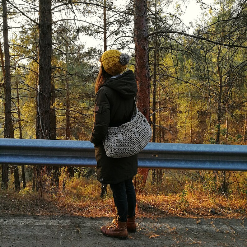 A model is stood at the side of the road looking out into the woods. She carries the mushroom bag across her body with the bulk of the bag behind her. It is full and you can clearly see the 3 crocheted mushrooms in a line in the middle of the bag.