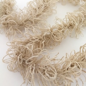 Puede incluir: Primer plano de una guirnalda de crochet beige hecha de cuerda trenzada. La guirnalda está hecha de una serie de bucles y nudos, creando una apariencia texturizada y esponjosa.