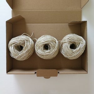 A birds eye view of three balls of beige hemp yarn sat horizontally in an open brown card box. The background is white.