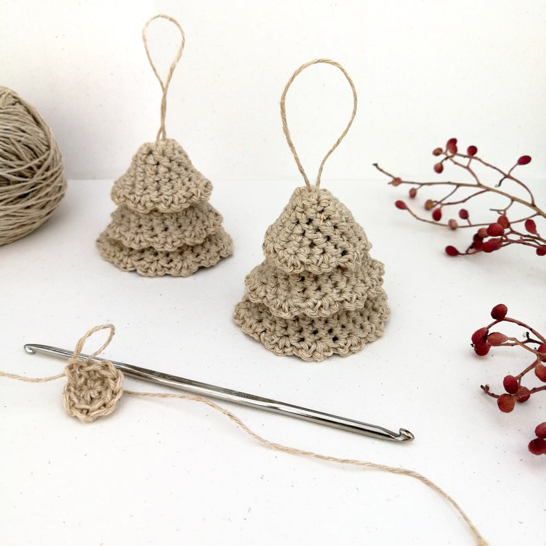 A couple of Christmas tree shaped ornaments are sat on a white background. They have three tiered skirts and a loop for them to be hung on the tree. A crochet hook is laid before them with the beginning of the next ornament near it.