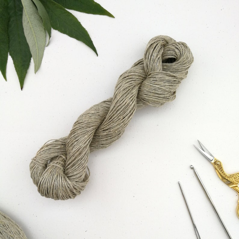 A skein of sport weight hemp yarn is laying diagonally on an off white background. The skein is light warm grey in colour and each strand is clearly defined. There is some green foliage in the top left corner and crochet tools in the bottom right.