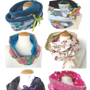 May include: Collection of six colorful Lupi Loop scarves displayed on mannequin heads. The scarves feature various patterns, including floral, comic book, and geometric designs. Each scarf is a different color, with some having ribbon accents.