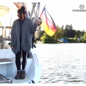 May include: A woman wearing a gray cardigan, black leggings with white stripes, and brown boots stands on a boat. A German flag is waving in the background.