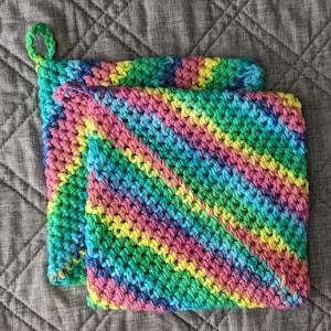 May include: Two crocheted pot holders in a rainbow color pattern. The pot holders are made with a green, blue, pink, and yellow yarn.