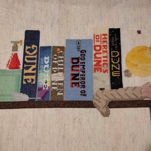 May include: A cross-stitch artwork depicting a bookshelf with a collection of books from the Dune series by Frank Herbert. The books are arranged in order of publication, with the first book, "Dune", on the bottom and the latest book, "Heretics of Dune", on the top. The bookshelf is decorated with a small, green plant and a red bottle. A cat is lying on top of the books.