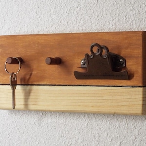 May include: A wooden key holder with two hooks and a metal clip. The wood is stained in two tones, a darker brown on top and a lighter brown on the bottom. A single key is hanging on one of the hooks.