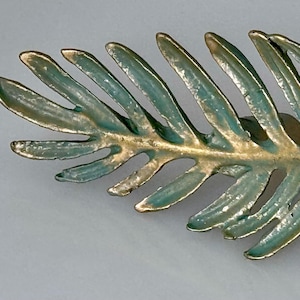 May include: A decorative leaf-shaped brooch with a gold-colored stem and edges. The leaf sections are a teal color, creating a two-tone effect. The brooch is likely made of metal and has a pin on the back.