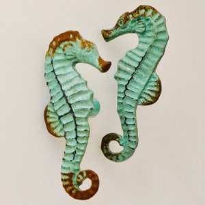 May include: Two decorative seahorse cabinet pulls. The seahorses are a distressed turquoise color with brown accents, giving them a rustic, aged appearance. The pulls are made of metal and are designed to be mounted on a cabinet or drawer.