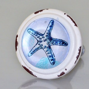 May include: A white cabinet knob with a distressed finish, featuring a blue starfish design set within a clear, round glass. The knob has a metal stem and is suitable for coastal or nautical decor.