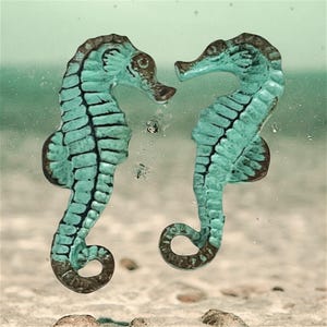 May include: Two seahorse-shaped ornaments featuring a textured, oxidized turquoise and brown finish. The seahorses are positioned facing each other, displaying detailed ridges along their bodies, curved tails, and prominent fins on their backs.