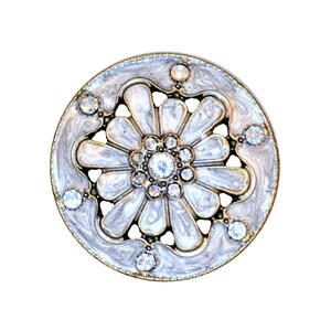 May include: A round, ornate brooch with a floral design. The brooch features a central cluster of clear crystals surrounded by white enamel petals and smaller crystals. The outer edge has a gold-tone metal border.