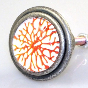 May include: A decorative cabinet knob with a silver-toned metal finish. The knob features a circular design with a white background and a vibrant orange, coral-like pattern. The knob is attached to a metal rod with a nut.