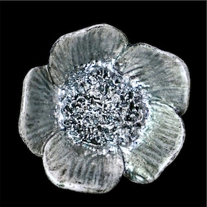 May include: A silver-toned flower-shaped object with a textured center and petals. The petals have a slightly rough, metallic appearance, and the center has a granular texture. The flower is set against a solid black background.