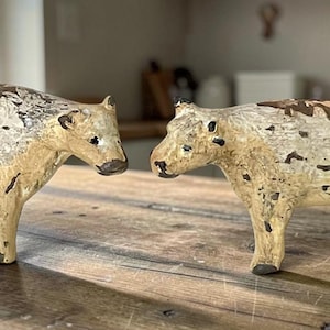 May include: Two standing cow figurines crafted with a distressed finish. The figures feature a cream-colored base with patches of dark brown paint, creating a weathered appearance. Each cow has four legs, a defined snout, and small ears.