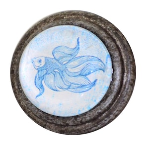 May include: Round decorative cabinet knob with a blue fish design. The fish is drawn in a whimsical style with flowing lines, set against a white and blue speckled background. The knob has a dark, textured, metallic frame.