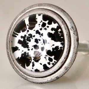 May include: A round cabinet knob with a black and white spotted design set within a distressed silver-colored metal frame. The knob has a glass-like appearance and is likely used for furniture or decorative purposes.