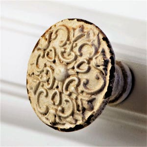 May include: Close-up of a distressed, off-white cabinet knob with an ornate, swirling design. The knob has a small, central protrusion and a slightly rusted, aged appearance, suggesting a vintage or antique style.