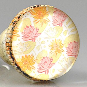 May include: A decorative cabinet knob with a floral design. The knob features a clear, round glass top with a pattern of orange and pink flowers and yellow lily pads on a white background. The sides have a textured, tan and brown pattern.