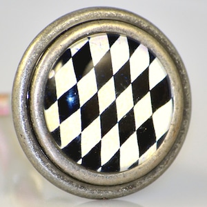 May include: Close-up of a round cabinet knob with a black and white checkered design. The knob has a silver-colored metal frame with a slightly distressed finish. The checkered pattern is in a diamond shape.