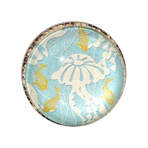 May include: Round, decorative glass cabochon with a jellyfish and fish design. The cabochon features a light blue background with a white jellyfish and yellow fish. The edge is framed with a silver-colored metal.