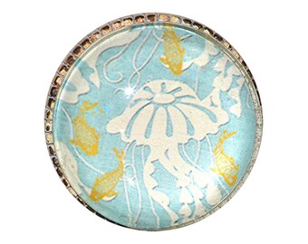 May include: Round, decorative glass cabochon with a jellyfish and fish design. The cabochon features a light blue background with a white jellyfish and yellow fish. The edge is framed with a silver-colored metal.