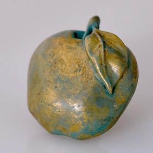 May include: A decorative apple sculpture with a metallic finish. The apple is primarily gold with patches of teal and a single leaf. The sculpture is a unique piece of home decor.
