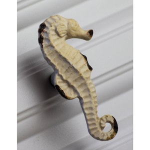 May include: Cream-colored seahorse-shaped cabinet knob with a distressed finish. The knob has a detailed design, with a curved tail and a textured surface. The seahorse is mounted on a white surface.