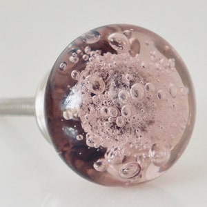 May include: A close-up of a round, clear glass cabinet knob with a silver metal stem. The knob is filled with a light pink, textured material and numerous air bubbles. The knob is a decorative hardware element.