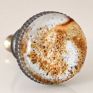 May include: A decorative cabinet knob with a round, textured face featuring a white and brown pattern, resembling coffee foam. The knob has a silver-colored metal base and a ridged edge, adding a touch of elegance to any furniture piece.