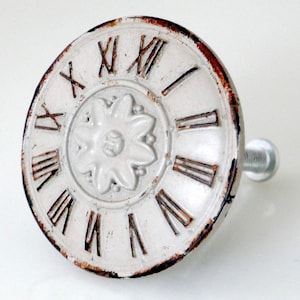 May include: A distressed white cabinet knob with a clock face design. The knob features Roman numerals and a central floral embellishment. The edges have a rusty brown finish, giving it a vintage look.