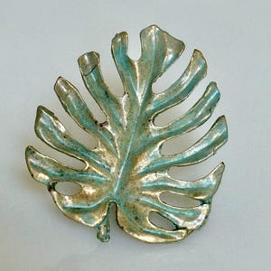 May include: A decorative leaf-shaped object with a metallic finish. The leaf has a detailed design with cut-out sections and a textured surface. The color is a blend of gold and teal, creating a vintage or antique appearance. The object is likely a brooch or decorative element.
