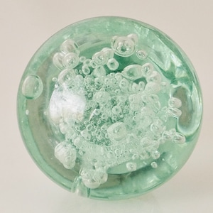 May include: A round, clear glass paperweight with a pale green tint. The interior is filled with numerous small, spherical air bubbles, creating a textured, bubbly appearance. The paperweight is smooth and rounded.