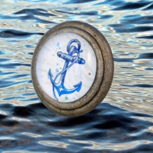 May include: A circular cabinet knob featuring a metallic bronze-toned frame and a white center insert. The insert displays a blue watercolor-style illustration of a nautical anchor surrounded by small blue paint splatters and dots.