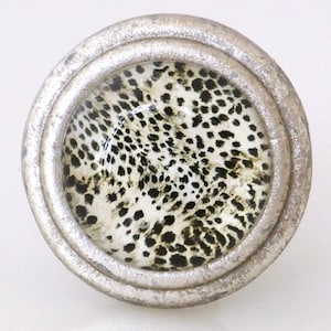 May include: A round, silver-toned cabinet knob with a glass insert featuring a black and white animal print pattern. The knob has a textured, metallic finish and a classic design, suitable for furniture or decor.