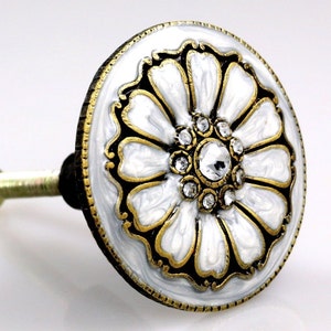 May include: Decorative white and gold floral cabinet knob with a central crystal and smaller surrounding crystals. The knob has a black border and a gold-colored metal stem.