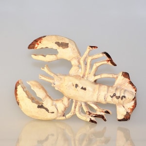 May include: A cream-colored lobster figurine with red-brown accents on the claws and tips. The lobster has a distressed, vintage appearance, with dark spots and a textured surface. The claws are open, and the figurine is set against a neutral background.