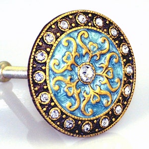 May include: Decorative cabinet knob with a round, ornate design. The knob features a light blue center with gold filigree and a border of clear crystals and dark bronze. A central crystal adds sparkle.